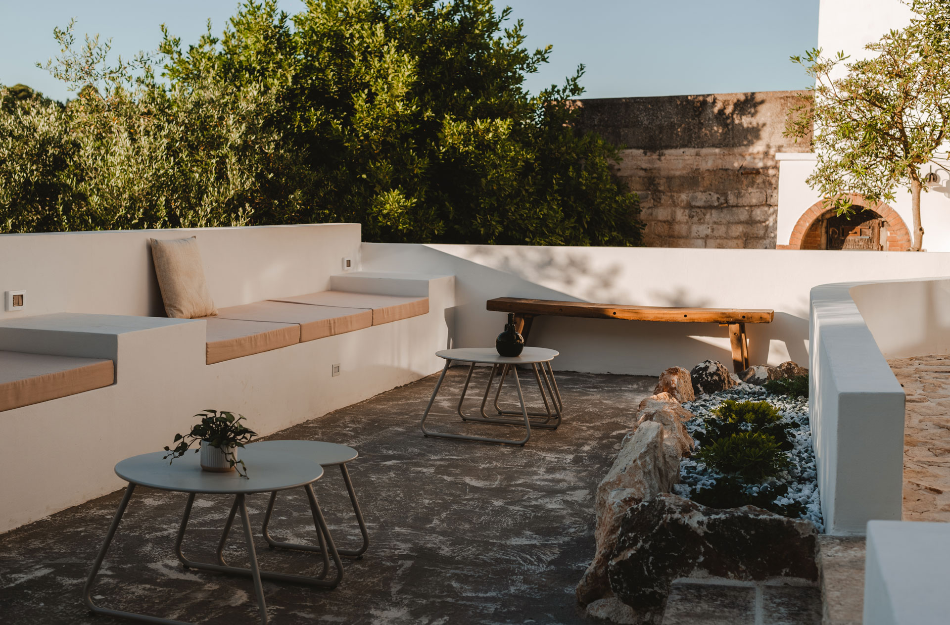 Terrazza con panche in muratura bianca, cuscini color sabbia, tavolini metallici, aiuola di rocce e muro in pietra di Dimora Maseva