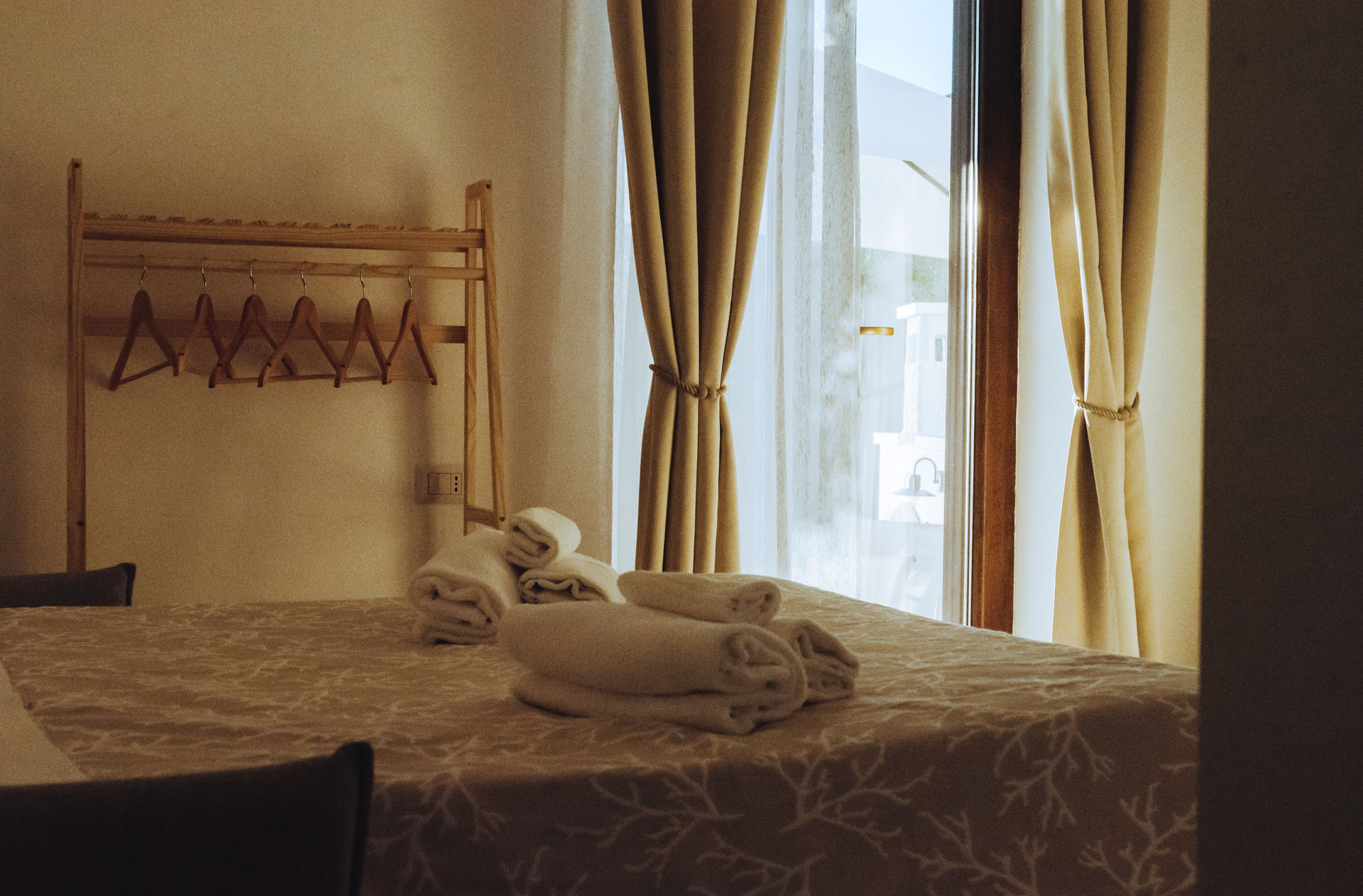 Camera di Dimora Maseva con asciugamani bianchi arrotolati sul letto, appendiabiti in legno e finestra con tende beige