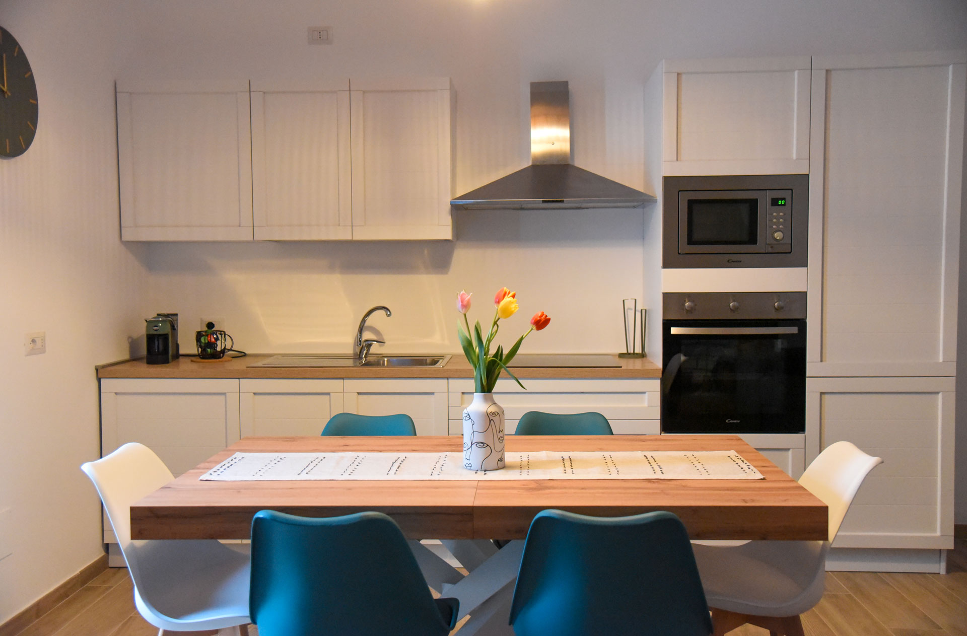 Cucina di Dimora Maseva con mobili bianchi, forno, microonde e tavolo in legno con tulipani e sedie bianche e blu