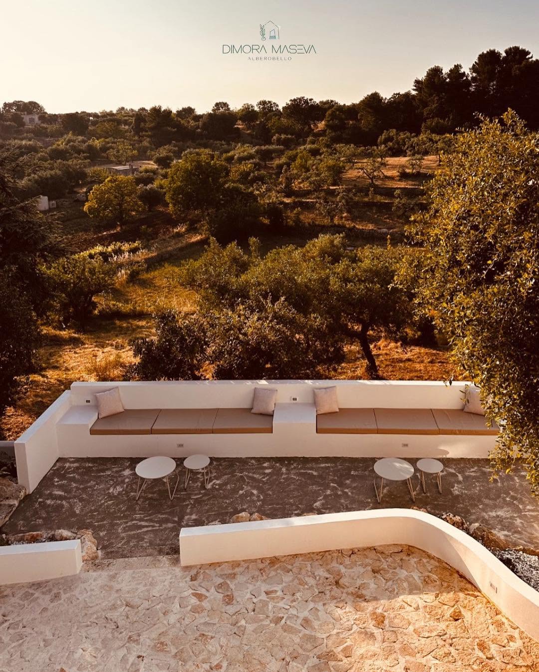 Zona chill out di Dimora Maseva: panche bianche in muratura, cuscini beige, tavolini tondi e ulivi al tramonto